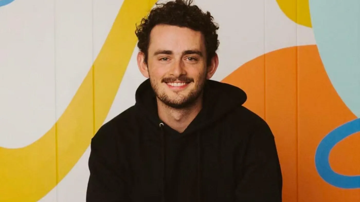 A smiling individual wearing a black hoodie sits in front of a colorful background featuring abstract shapes in yellow, orange, and blue. A smiling individual wearing a black hoodie sits in front of a colorful background featuring abstract shapes in yellow, orange, and blue.
